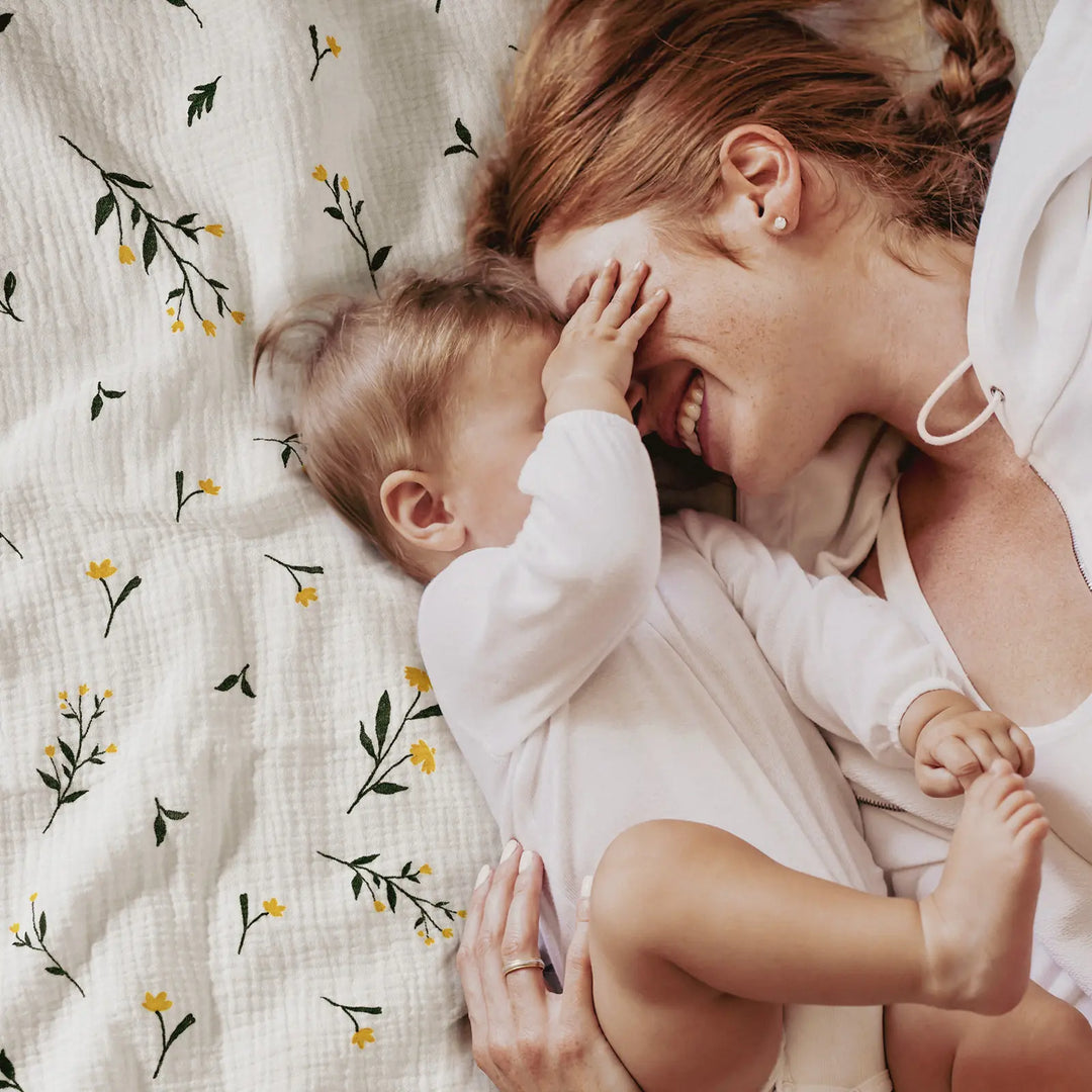 A mother and her baby cuddling on a floral baby blanket, both smiling and sharing a playful moment.