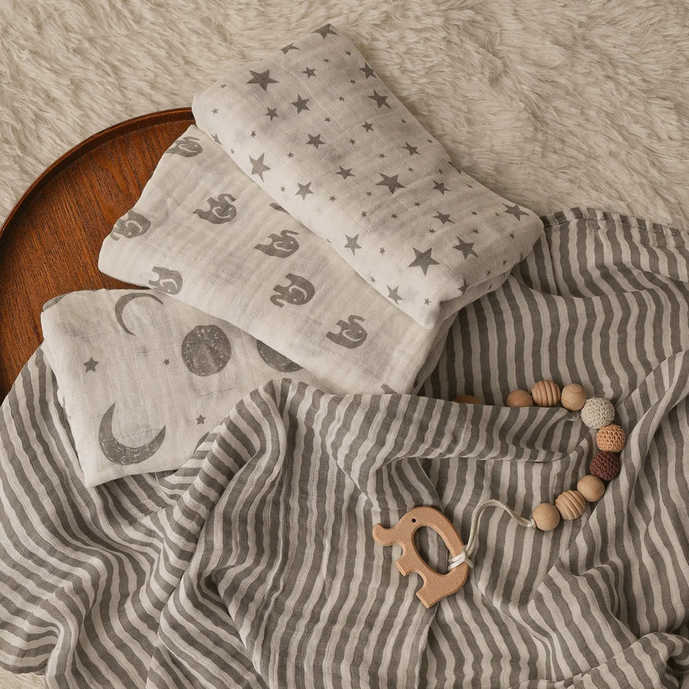 Set of baby muslin swaddle blankets with various patterns, including elephants, moons, stars, and stripes, displayed on a cozy surface with a wooden elephant teether and a beaded pacifier clip. Perfect for newborns and infant essentials.