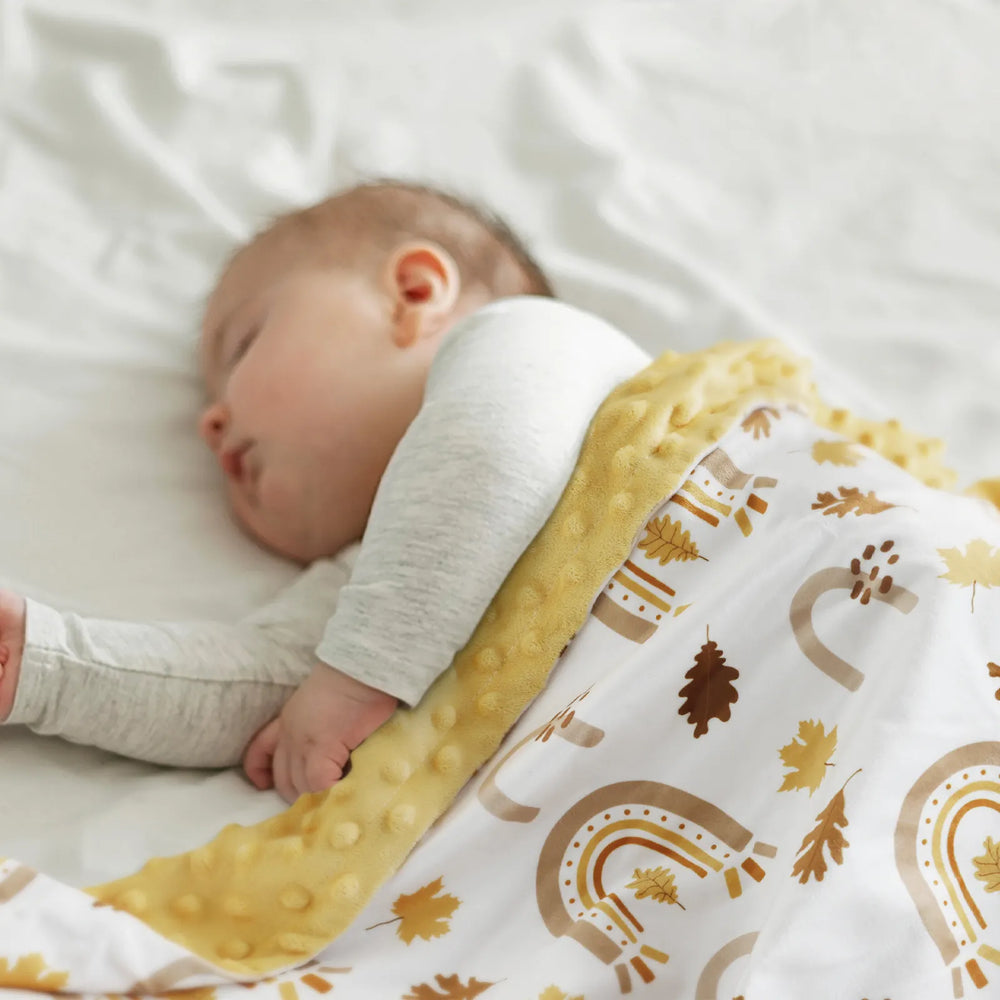 Baby peacefully sleeping under a cozy fall-themed minky blanket with a rainbow and leaf pattern, featuring a soft yellow reverse side.