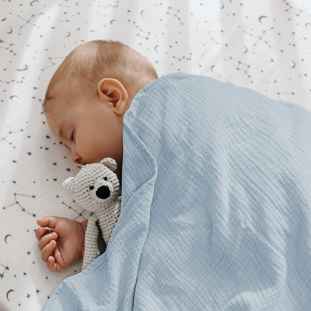 Sleeping baby wrapped in light blue muslin swaddle blanket with knitted teddy bear on star-patterned background, demonstrating blanket's comfort