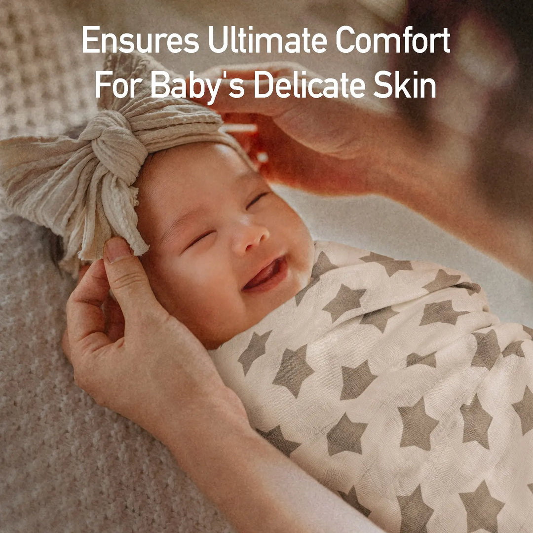 Smiling baby wrapped in a soft star-pattern muslin swaddle blanket, with a bow headband. Text overlay reads 'Ensures Ultimate Comfort For Baby's Delicate Skin.