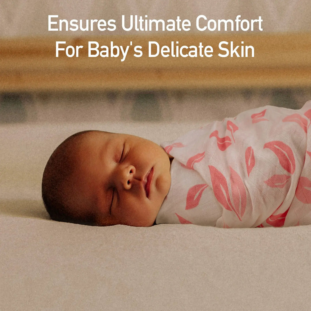 Newborn baby peacefully sleeping wrapped in a pink leaf-pattern muslin swaddle blanket with text overlay reading 'Ensures Ultimate Comfort For Baby's Delicate Skin.