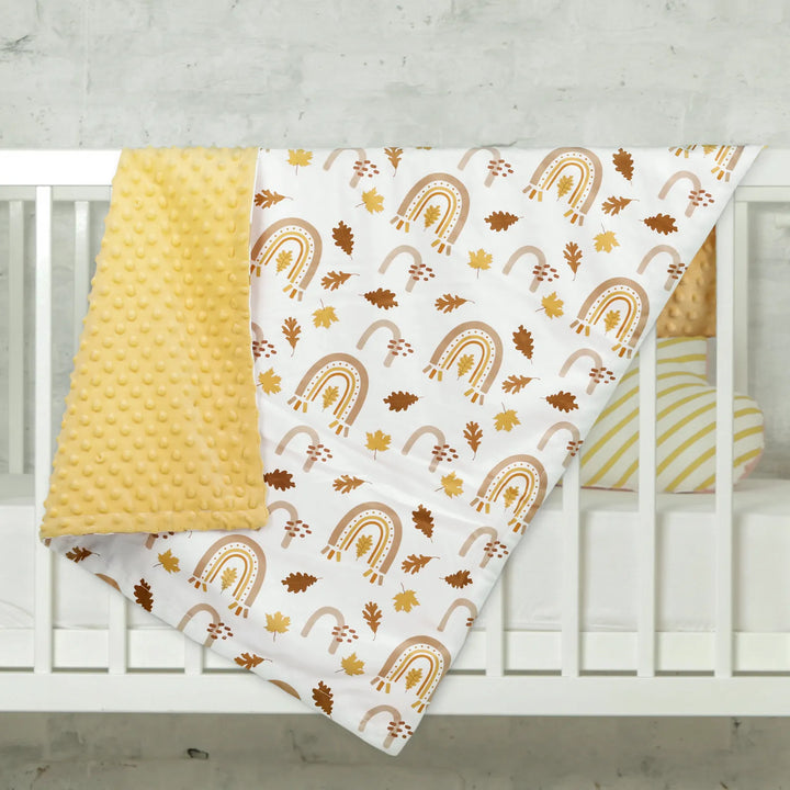 Fall-themed minky blanket with rainbow and autumn leaves pattern draped over a crib, showing a soft yellow reverse side. Ideal for a cozy nursery setting.