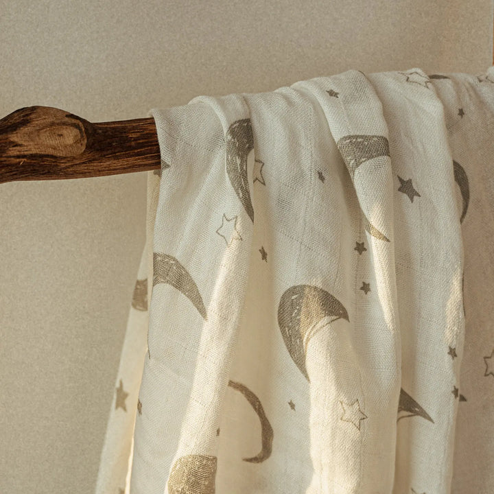 Close-up of a muslin baby swaddle blanket with a delicate moon and stars pattern, draped over a wooden rod. Soft, breathable fabric ideal for newborns and infants.