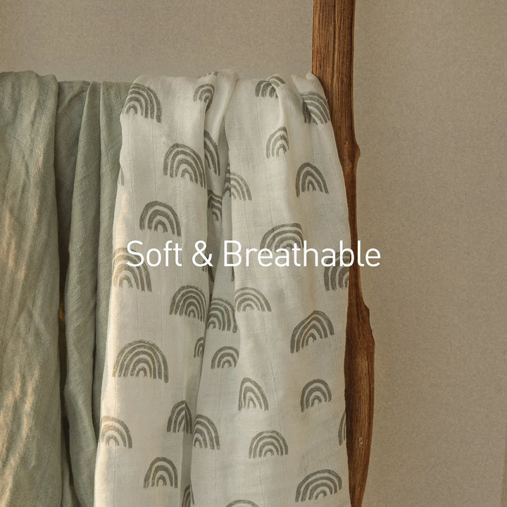 Neutral muslin swaddle blankets with rainbow print and solid colors, draped over a wooden ladder. Text overlay reads 'Soft & Breathable.' Perfect for newborns.