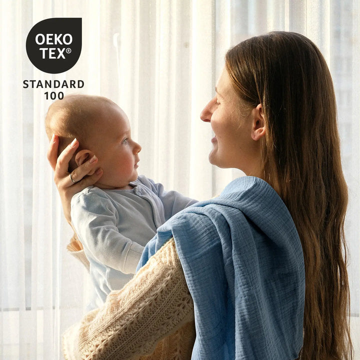 Mother holding baby with a blue Organic Cotton Muslin Swaddle Blanket, OEKO-TEX® Standard 100 certified for safety and softness.