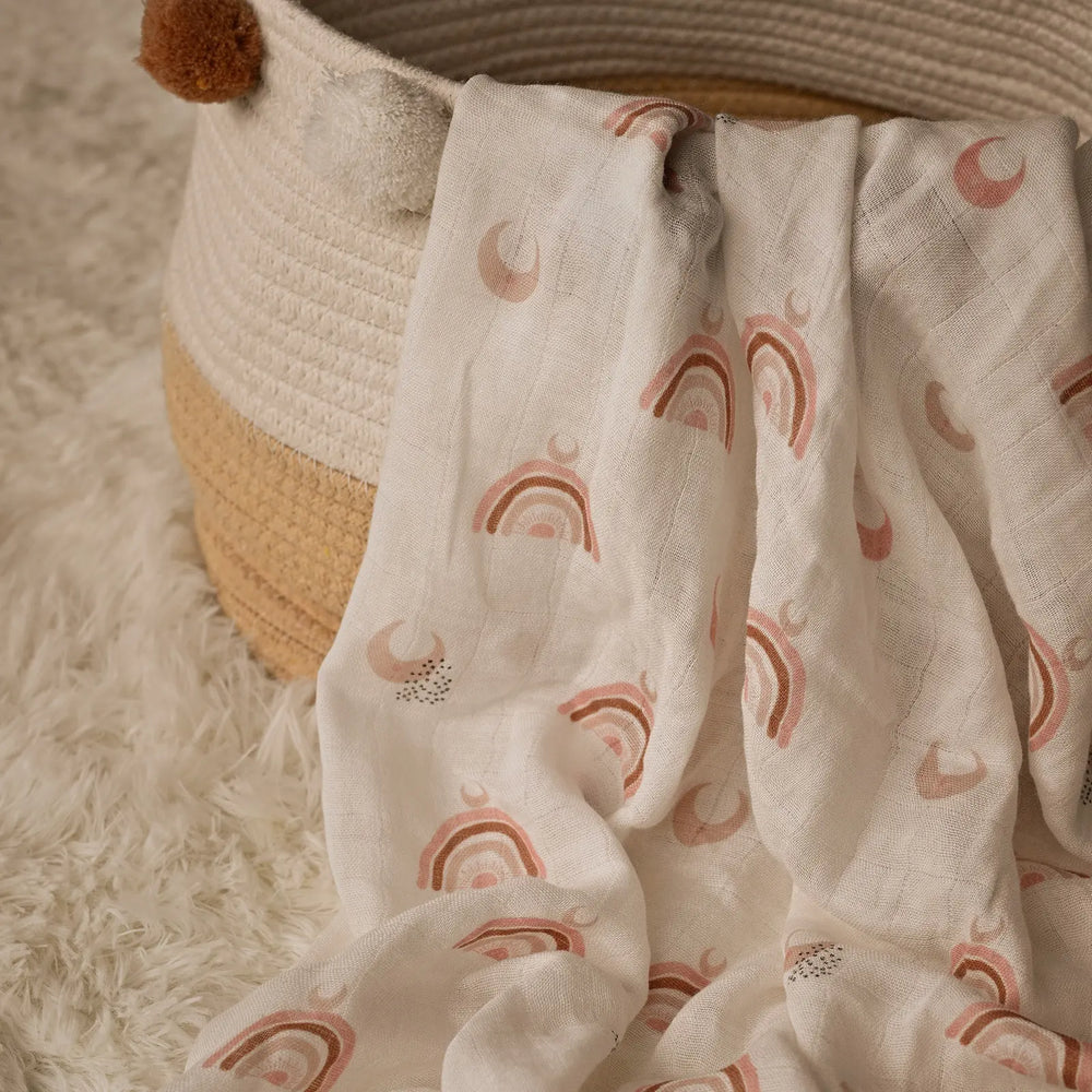 Muslin swaddle blanket with warm-toned rainbow pattern draped over a woven basket on a soft, textured surface. Ideal for creating a cozy nursery atmosphere.