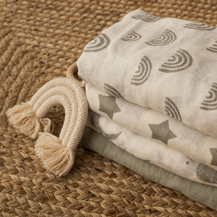 Stack of neutral-themed muslin swaddle blankets with rainbow, star, and moon patterns, accented with a woven tassel rainbow. Cozy and stylish for babies.