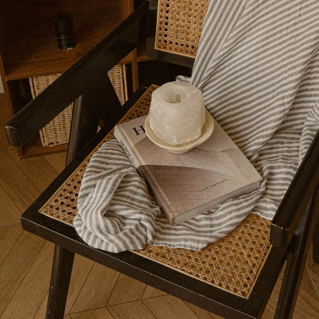 Cozy aesthetic scene featuring a striped blanket draped over a woven cane chair, with a decorative book and a rustic candle placed on top. Perfect for home decor inspiration.